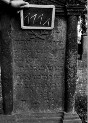 Grabstein 111 (Index: 100111230) Material: Sandstein Zustand: verwittert Maße HxBxT: 145x70x19 cm Beschreibung: ädikulaartige Stele mit Halbsäulen, getreppter Sockel, giebelartiger Abschluß mit Verdachung Symbolik: gekreuzte Zweige, Blüte, Zackenfries Besonderheit: Kindergrab: Grabstein 111A