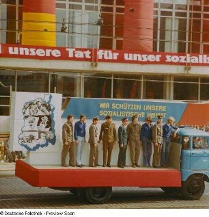 Demonstrationszug zum 1. Mai. Teilnehmer (uniformiert und in FDJ-Kleidung) auf LKW mit der Losung "Wie schützen unsere sozialistische Heimat