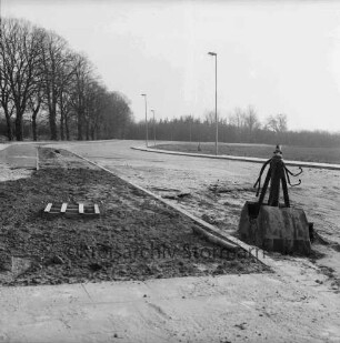 Gewerbegebiet West: Straßenbau: vorn unbefestigtes Gelände, rechts Baggerschaufel: hinten Straße, Fußwege und Straßenlampen: dahinter Bäume