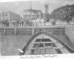 Barbados. Hafenszene mit Einheimischen und Touristen auf der Kaimauer, im Hintergrund eine Kirche. Blick aus einem vertäuten Ruderboot