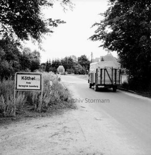 Köthel (Kreis Herzogtum Lauenburg): Ortseingang mit landwirtschaftlichem Fuhrwerk: links Ortseingangsschild: im Hintergrund Ehrenmal beider Orte am Billehof: hinten Bäume