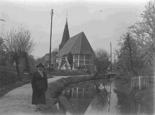 Mit Friedhof umstandene Fachwerk-Kirche neben einem Wasserlauf und einer Frau im Vordergrund