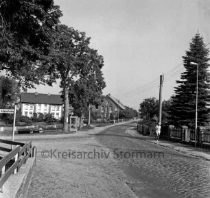Dorfstraße mit Kopfsteinpflaster: links Abzweigung der Lindenstraße: davor Zaun: dahinter Haus und Bäume: rechts Zaun: davor Jogger auf Fußgängerweg