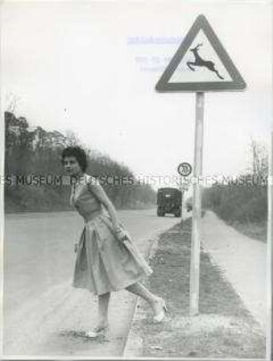 Vor einem Verkehrsschild "Wildwechsel Rehe" überquert ein "rehähnlich" graziles Mädchen die Straße (ironische Fotografie)
