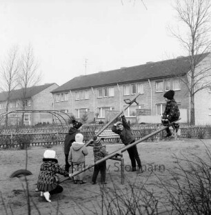 Kinderspielplatz für Siedlung Gartenstadt: Kieler Straße: Kinder auf den Wippen: dahinter Klettergerüst: Jägerzäune, Sitzbank, Baum: Fußweg: hinten Gartenfront der Reihenhäuser an Jahnstraße