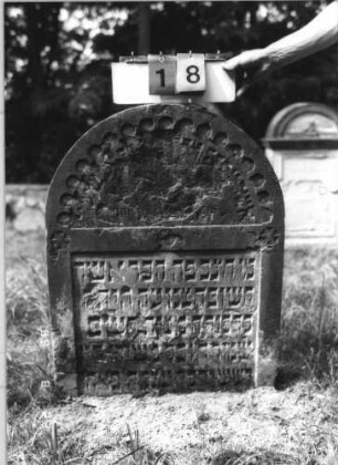 Grabstein 18 (Index: 100110924) Material: Sandstein Zustand: im Giebel Abschieferungen mit großem Textverlust, im unteren Teil leicht verwittert mit geringem Textverlust, eingesunken Maße HxBxT: 95x73x11 cm Beschreibung: Stele, Rundbogenabschluß mit Rundbogenfries, Schriffelder eingetieft Symbolik: 3 Rosetten: Grabstein 18