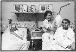 Krankenschwester schüttelt das Kissen eines Kranken auf, der in seinem Bett sitzt, ein zweiter Mann liegt im Bett auf der anderen Seite und schläft (Altersgruppe 18-21)