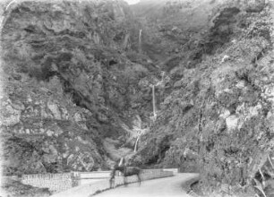 Straße : Eine Straße, die an Felsen und einem Wasserfall entlang führt. Ein Mann steht an der Straße mit einem Pferd