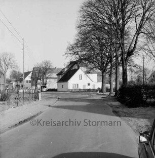 Hauptstraße: rechts Baumreihe hinter Zaun und Hecke: hinten Wohn- und Geschäftshäuser: davor parkende Pkws: links Zaun und Strommast: dahinter Garten