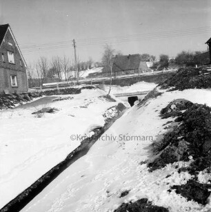 Mühlenbach: stillgelegter Ausbau: rechts Erdhaufen: hinten rechts Durchlass unter Straße: links und im Hintergrund Strommast, Häuser und Bäume: Winter