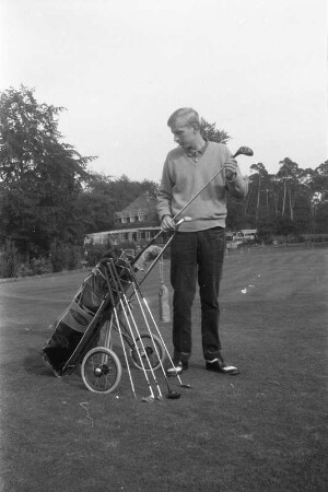 Junger Golfspieler neben Golftrolley mit Golfbag und Golfschlägersatz, aufgenommen auf einem Golfplatz