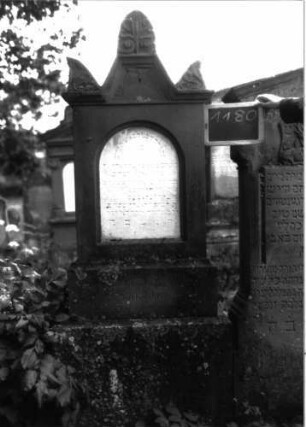 Grabstein 1180 (Index: 100104324) Material: Sandstein, Marmortafel Zustand: gut erhalten, im Giebel beginnende Verwitterung Maße HxBxT: 198x57x22 cm Beschreibung: Stele auf getrepptem Sockel, giebelartiger, hochgezogener Abschluss mit Eck- und Giebelakroterien (Voluten- u. Blattornamente), eingetieftes, rundbogiges Schriftfeld Symbolik: Blattornamente: Grabstein 1180