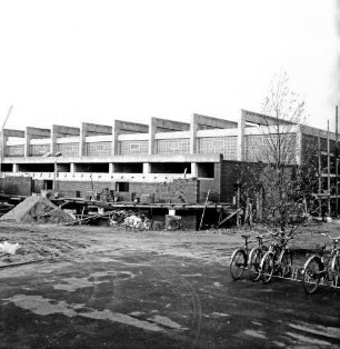 Volksschule: Sporthalle: Rohbau: davor Gerüste, Baumaterial und Sandhaufen: vorn rechts Fahrradständer mit Rädern: dahinter Baum