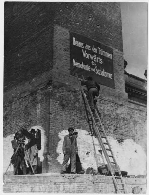 Postament des Denkmals für Friedrich den Großen mit Losung "Heraus aus den Trümmern - Vorwärts für Demokratie und Sozialismus". Szene mit Kamerateam nach dem Anbringen des Plakates