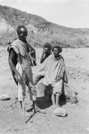 Gruppe der Arsi-Oromo : Ein älterer Mann mit Stock in der Hand steht vor einem Stein auf dem drei Personen sitzen