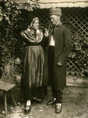 Ehepaar stehend, Frau gestützt an historischem Holzstuhl, Mann rechts, der Ehefrau zugewandt mit Pfeife. Halbtotale