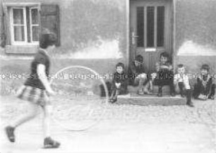 Mädchen mit einem Hula-Hoop Reifen und Kindergruppe vor Haustür eines alten Hauses