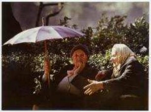 Zwei alte Frauen auf einer Parkbank, eine im Schatten ihres Regenschirms, die andere, mit Kopftuch, sitzt in der Sonne