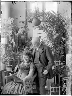 Wohnung der Familie Knochenhauer, Wintergarten? Dame, mit einem Buch im Korbstuhl sitzend, dahinter Herr (Ehemann?) stehend : Kattowitz, Friedensstraße 2. Wohnung der Familie Knochenhauer, Wintergarten? Dame, mit einem Buch im Korbstuhl sitzend, dahinter Herr (Ehemann?) stehend