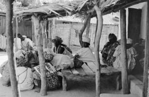 Gruppe von Frauen in einem Innenhof mit strohgedeckten Lehmhütten bei einer traditionellen Initiationszeremonie, Gambia