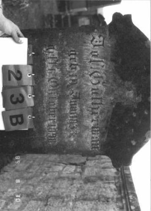 Grabstein 23 (Index: 99609659) Material: Sandstein Zustand: verwittert Maße HxBxT: 189x63x24 cm Beschreibung: Stele, Segmentbogenabschluß, in Voluten auslaufend, Volutenaufsatz, Blüte, Schriftfeld eingetieft, getreppter Sockel Symbolik: Kranz, Blüte: Grabstein 23B