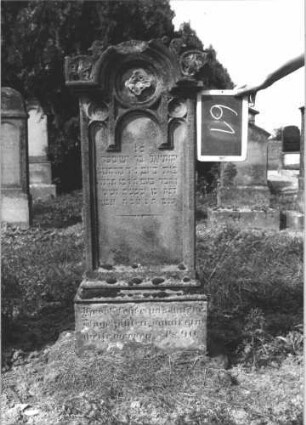 Grabstein 61 (Index: 100114035) Material: Sandstein Zustand: verwittert Maße HxBxT: 120x52x19 cm Beschreibung: Stele, Sockel, eingezogener Rundbogenabschluß mit Verdachung und Eckakroteren, Schriftfeld mit drei Rundbögen Symbolik: Blüten, stilisierte Blätter: Grabstein 61