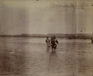 Durchqueren des Tirgirs : Drei Personen durchqueren den Tigris. Ein Mann trägt ein Kind auf dem Rücken durch den Fluss.