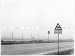 Eine Straße mit Schild, das auf Flugzeuge hinweist, dahinter ein Zaun mit Stacheldraht, weit hinten ein Bulldozer (Altersgruppe 14-17)