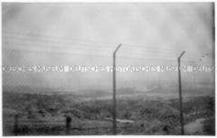 Durch einen zweiten Zaun hindurch fotografierte, verschwommene Aufnahme eines Maschendrahtzauns mit Stacheldraht, dahinter die freie Fläche einer Baustelle (Altersgruppe 14-17)