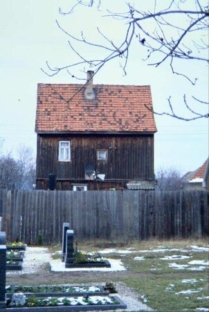 hohes schmales Haus mit Bretterverkleidung; vorn Friedhof