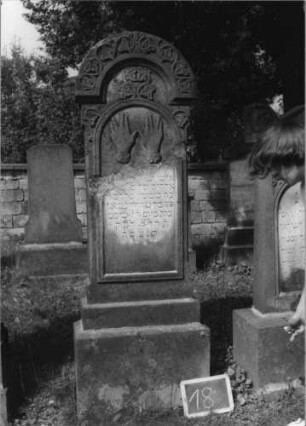 Grabstein 18 (Index: 99801905) Material: Sandstein Zustand: gebrochen und repariert mit Textverlust Maße HxBxT: 190x50x20 cm Beschreibung: Stele, getreppter Sockel, Rundbogen, eingetieftes Schriftfeld mit Rundbogen Symbolik: segnende Hände, Blätterfries: Grabstein 18