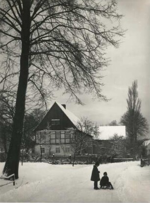 Verschneites Haus mit Bäumen und spielenden Kindern mit Schlitten (vielleicht Seiffen-Heidelberg oder Ullersdorf bei Dresden)