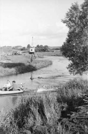 Trave: Flussregulierung: vorn Uferböschung mit Schilf: rechts Baum: links Motorboot mit zwei Personen im Wasser: dahinter Mann, Bagger, Loren und Baumaterial: hinten Wiesen und Büsche: dahinter Häuser von Sehmsdorf zwischen Bäumen