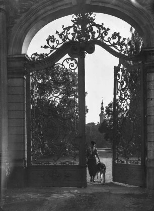 Unbekannter Ort. Junge Frau mit Hund an einem schmiedeeisernen Gittertor stehend, Blick zu einem Kirch- oder Schlossturm