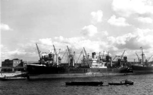 Hamburg. Im Hafen liegen Frachtschiffe an den Kaianlagen. Im Vordergrund fährt ein Schlepper mit einer Schute