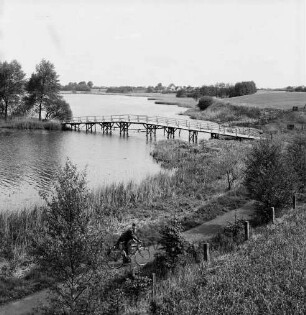 Herrenteich: Fischhuser Damm: hölzerne Fußgängerbrücke: Blick Richtung Nordosten auf oberen Herrenteich: im Vordergrund Ufer mit Schilf, Büsche, Zaun, Wanderweg, Fahrradfahrer