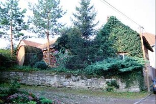 Natursteinmauer, darüber Hausgiebel mit Begrünung und großes Fass