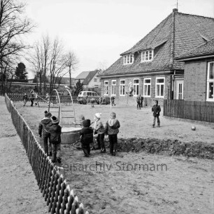 Kindergarten in ehemaligem Volksschulgebäude: Schulstraße: im Vordergrund Hof mit Spielgerät und spielenden Kindern: links Jägerzaun: hinten Pkw, Bäume und Wohnhäuser