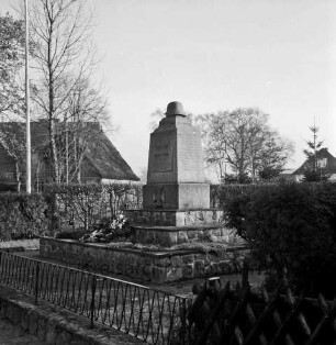 Dorfstraße: Ehrenmal mit Kranz- und Blumenschmuck: vorn Zaun: hinten Hecke: hinten Häuser und Bäume