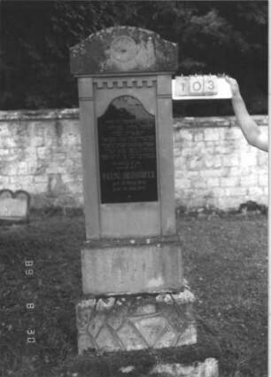 Grabstein 103 (Index: 99609739) Material: Sandstein, Inschriftplatte Glas Zustand: leicht verwittert, Inschriftplatte gesprungen Maße HxBxT: 192x62x26 cm Beschreibung: Stele, schräg eingezogener flacher Segmentbogenabschluß, eingelassene Schriftplatte mit Konvexbogenabschluß, getreppter Sockel, unten rusticaartig: Grabstein 103