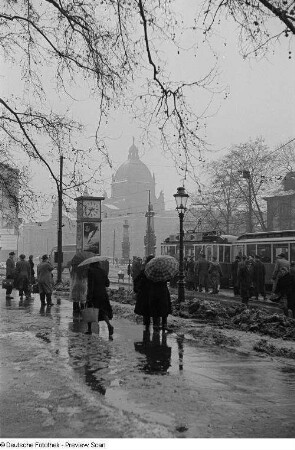 Menschen mit Regenschirmen warten an der Straßenbahnhaltestelle auf dem Martin-Luther-Ring, im Hintergrund das Reichsgericht