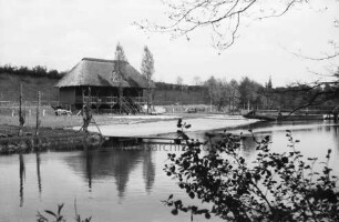 Trave: hinten links Sportlerhaus: davor Sandstrand: links Steg: hinten rechts Holzbrücke und Boot am Ufer: dahinter Bäume, Sportplatz und Hang am Trave-Stadion: vorn rechts Baumäste