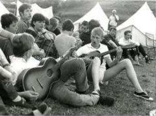 Jugendliche sitzen vor Zelten und singen, zwei mit Gitarren