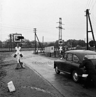 Siebenbäumen (Kreis Herzogtum Lauenburg): Bahnstrecke zwischen Bad Oldesloe und Ratzeburg: Bahnübergang der Landstraße zwischen Bad Oldesloe und Ratzeburg: Filmaufnahmen der Reginald Puhl Filmproduktion aus Hamburg-Wandsbek zum Dokumentarfilm "Todesfallen": nachgestellter Bahnunfall einer Dampflokomotive mit einem Pkw: in der Mitte Bahnübergang mit Signalanlagen: vorn rechts Mann beim Schieben des Pkws: dahinter Strommasten: hinten rechts Dampflokomotive sowie Häuser hinter Bäumen: im Hintergrund Wiesen