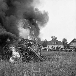 Dorffest: Brandmanöver: brennende Holzstapel (vorne) und brennende Holzhütte dahinter, im Hintergrund Zuschauer