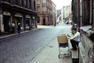 DDR. Straßenbild mit jungem Mann, neben einem Kinderwagen vor einem Hauseingang sitzend, gegenüber das HO-Kaufhaus West