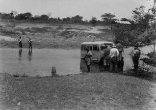 Flussüberquerung : Der Expeditionsbus durchquert einen Fluss an einer seichten Stelle. Die Mitglieder der Expedition stehen um den Bus versammelt. Möglichweise steckt dieser im Flussbett fest.