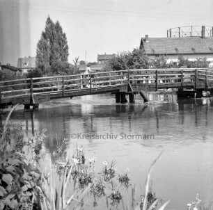 Trave: Hochwasser: vorn links Grünpflanzen: in der Mitte Kranbrücke: darauf Frau mit Fahrrad: hinten rechts Gebäude der Hamburger Tapetenfabrik: dahinter Gasometer der Stadtwerke: hinten links hinter Bäumen Häuser an der Lübecker Straße