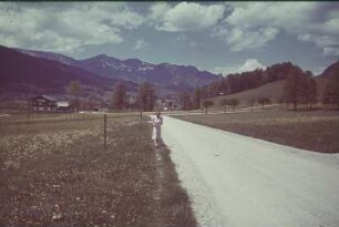 Deutschland. Junge Frau an einer Landstraße vor einer Bergkette (vielleicht in Bayern)