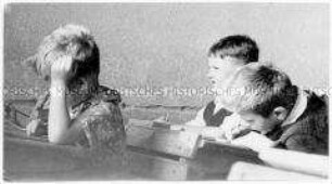 Drei Jungen sitzen in Schulbänken, einer schreibt